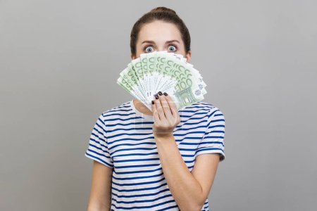 Photo for Portrait of surprised woman wearing striped T-shirt peeping out euro banknotes with exited look, big jackpot, financial success. Indoor studio shot isolated on gray background. - Royalty Free Image