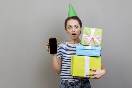 Photo for Portrait of shocked woman wearing striped T-shirt and party cone, holding stack of present boxes and showing mobile phone with blank display. Indoor studio shot isolated on gray background. - Royalty Free Image