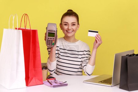 Photo for Smiling satisfied woman office employee sitting at workplace with shopping bags, showing terminal and credit card to camera. Indoor studio studio shot isolated on yellow background. - Royalty Free Image