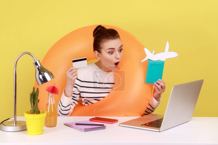 Photo for Amazed shocked woman with rubber ring holding and showing credit card and passport, looking at laptop display with open mouth. Indoor studio studio shot isolated on yellow background. - Royalty Free Image