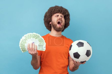 Photo for Excited man with Afro hairstyle wearing orange T-shirt holding soccer ball and hundred euro bills, screaming with happiness, betting and winning. Indoor studio shot isolated on blue background. - Royalty Free Image