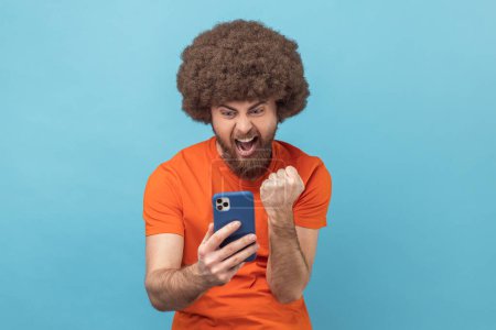 Téléchargez les photos : Portrait d'un homme extrêmement heureux avec une coiffure afro portant un t-shirt orange tenant un téléphone intelligent, regardant l'affichage, gagnant à la loterie, le poing serré. Studio intérieur tourné isolé sur fond bleu. - en image libre de droit