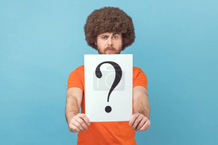 Photo for Portrait of man with Afro hairstyle wearing orange T-shirt holding question mark, finding smart solution, asking for advice, looking at camera. Indoor studio shot isolated on blue background. - Royalty Free Image