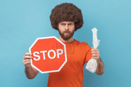 Photo for Portrait of serious responsible man with Afro hairstyle wearing orange T-shirt holding red stop sign and plastic package, looking at camera. Indoor studio shot isolated on blue background. - Royalty Free Image