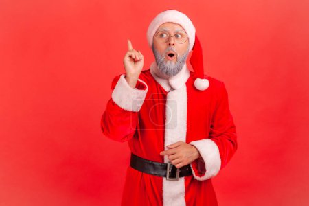 Photo for Elderly man with gray beard wearing santa claus costume raising finger and having genius idea of Christmas celebration, looking amazed, creative thought. Indoor studio shot isolated on red background. - Royalty Free Image