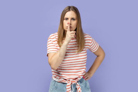 Photo for Shh, be quiet. Portrait of serious woman wearing striped T-shirt showing silence gesture with finger on her mouth, asking to stay quiet, keep secret. Indoor studio shot isolated on purple background. - Royalty Free Image