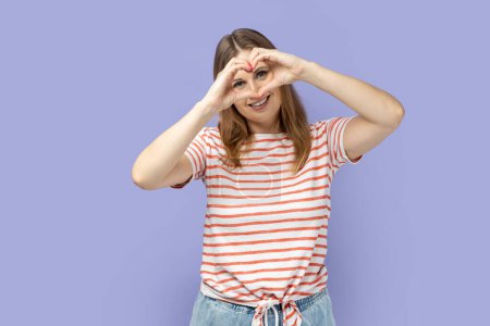 Photo for Portrait of blond woman wearing striped T-shirt making heart shape with hands in front of her eyes, expressing love, friendship, care and romance. Indoor studio shot isolated on purple background. - Royalty Free Image