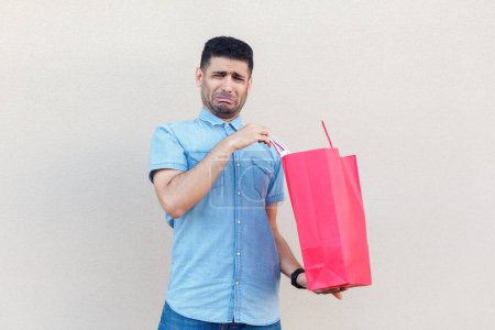Photo for Portrait of sad stressed depressed man wearing denim shirt being disappointed of his shopping in mall, holding paper bag and crying. Indoor studio shot isolated on gray background. - Royalty Free Image