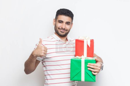 Photo for Portrait of attractive cheerful joyful bearded man wearing striped t-shirt standing looking at camera, holding two gift boxes, showing like gesture. Indoor studio shot isolated on gray background. - Royalty Free Image