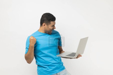 Téléchargez les photos : Joyeux homme joyeux non rasé en T-shirt bleu debout tenant l'ordinateur portable dans les mains, serre le poing, célèbre le succès, finissant le projet jusqu'à la date limite. Studio intérieur tourné isolé sur fond gris. - en image libre de droit