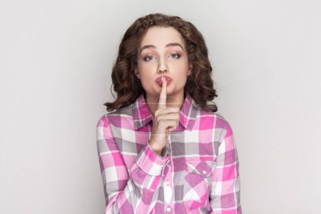 Photo for Pleasant looking delightful woman with curly hair keeps finger on lips, asks not tell secret information or keep silence, wearing pink checkered shirt. Indoor studio shot isolated on gray background. - Royalty Free Image
