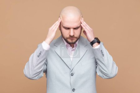 Photo for Portrait of sick ill unhealthy bald bearded man standing with hands on head, keeps eyes closed, suffering terrible headache, wearing gray jacket. Indoor studio shot isolated on brown background. - Royalty Free Image