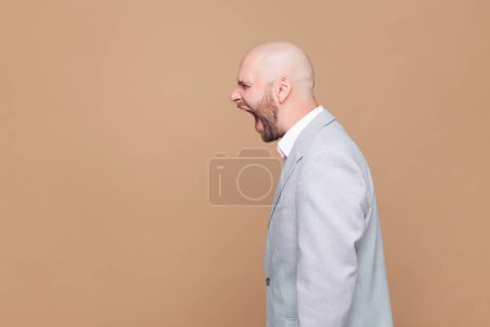 Photo for Side view portrait of angry or shocked bald bearded man standing, looking forward and screaming, copy space foe advertisement, wearing gray jacket. Indoor studio shot isolated on brown background. - Royalty Free Image