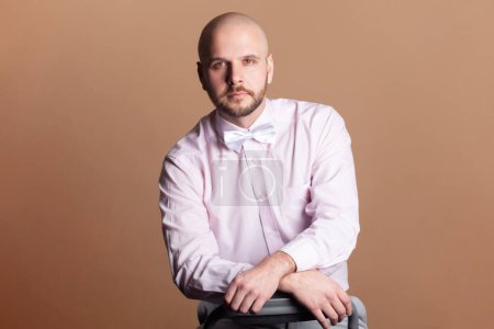 Photo for Portrait of handsome attractive bald bearded man looking at camera with serious concentrated facial expression, wearing light pink shirt and bow tie. Indoor studio shot isolated on brown background. - Royalty Free Image