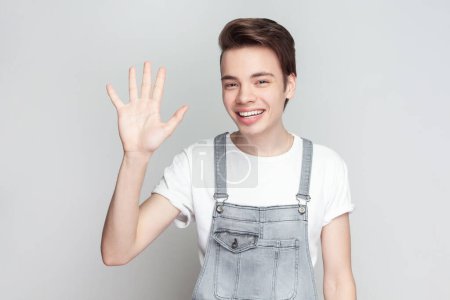 Photo for Cheerful friendly young brunette man standing waving palm in hello gesture, meets someone at street, smiles positively, wearing denim overalls. Indoor studio shot isolated on gray background. - Royalty Free Image