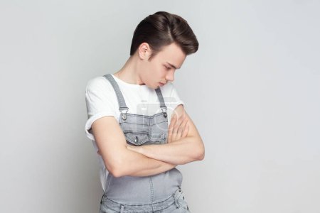 Photo for Portrait of sad unhappy young brunette man standing with folded hands, frowns face upset with bad news he received, wearing denim overalls. Indoor studio shot isolated on gray background. - Royalty Free Image
