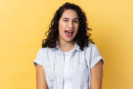 Photo for Portrait of cheerful attractive woman with dark wavy hair being in good mood, smiling broadly and winking at camera with toothy smile. Indoor studio shot isolated on yellow background. - Royalty Free Image