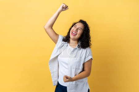 Photo for Portrait of excited happy woman with dark wavy hair expressing winning gesture with raised fists and screaming, celebrating victory. Indoor studio shot isolated on yellow background. - Royalty Free Image