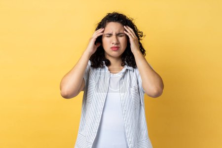 Photo for Portrait of woman with dark wavy hair frowning and clasping sore head, suffering intense headache, having migraine, fever symptoms. Indoor studio shot isolated on yellow background. - Royalty Free Image