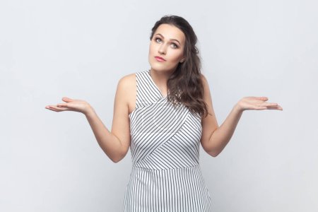 Photo for Portrait of brunette woman spreads palms, shrugs shoulders with perplexed expression, being indecisive, has no idea what happened, wearing striped dress. Indoor studio shot isolated on gray background - Royalty Free Image