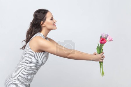 Téléchargez les photos : Portrait latéral de jolie femme brune tenant un bouquet de tulipes, le donnant à sa mère, félicitant le 8 mars, portant une robe rayée. Studio intérieur tourné isolé sur fond gris. - en image libre de droit