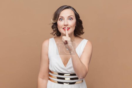 Photo for Portrait of woman makes silence or hush gesture, keeps index fingers over lips, tells secret information to someone, wearing white dress. Indoor studio shot isolated on light brown background. - Royalty Free Image