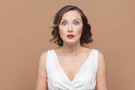 Photo for Portrait of shocked astonished middle aged woman with wavy hair looking at camera with big eyes, sees something excited, wearing white dress. Indoor studio shot isolated on light brown background. - Royalty Free Image