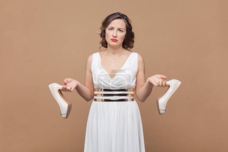 Photo for Portrait of sad upset middle aged woman with wavy hair holding elegant shoes on high heels, feels pain on feet, wearing white dress. Indoor studio shot isolated on light brown background. - Royalty Free Image