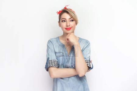 Photo for Portrait of positive smiling optimistic blonde woman wearing blue denim shirt and red headband standing looking away, dreaming. Indoor studio shot isolated on gray background. - Royalty Free Image