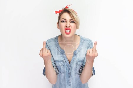 Téléchargez les photos : Portrait of angry aggressive rude blonde woman wearing blue denim shirt and red headband standing showing fuck sign, arguing with somebody. Indoor studio shot isolated on gray background. - en image libre de droit