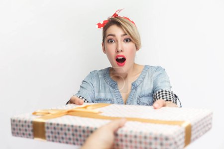 Photo for Portrait of astonished shocked blonde woman wearing blue denim shirt and red headband standing receives present box, being surprised. Indoor studio shot isolated on gray background. - Royalty Free Image