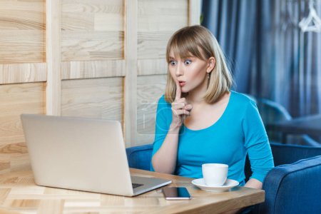 Photo for Portrait of serious adorable young woman with blonde hair in blue shirt sitting at table and working on laptop, has video call, showing shh gesture. Indoor shot, cafe background. - Royalty Free Image