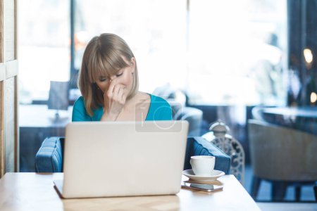 Photo for Portrait of sad despair upset young woman with blonde hair in blue shirt working on laptop, crying, hiding her face, having problems with her work. Indoor shot in cafe with big window on background. - Royalty Free Image