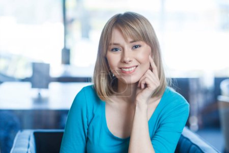 Téléchargez les photos : Portrait de souriante heureuse jeune femme satisfaite aux cheveux blonds en chemise bleue assise seule, regardant la caméra avec le sourire des dents, étant de bonne humeur. Intérieur tourné dans un café avec une grande fenêtre sur fond - en image libre de droit