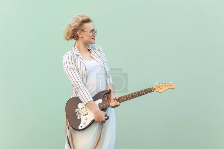 Photo for Portrait of beautiful positive blonde woman in white shirt, skirt, and striped blouse with eyeglasses, holding guitar and looking away. Indoor studio shot isolated on light green background. - Royalty Free Image