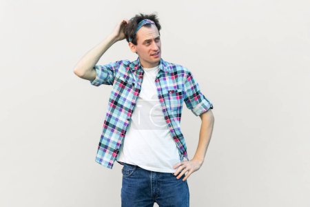Photo for Indignant puzzled man rubbing head, tries to remember necessary information, looks in displeasure, wearing blue checkered shirt and headband. Indoor studio shot isolated on gray background. - Royalty Free Image