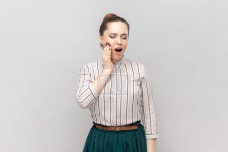Photo for Portrait of sick unhealthy ill woman wearing striped shirt and green skirt standing touching her painful cheek, suffering terrible toothache. Indoor studio shot isolated on gray background. - Royalty Free Image