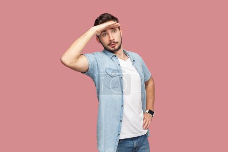 Photo for Bearded man in blue casual style shirt standing keeps palm near forehead, looks with concentrated expression into distance, sees something aside. Indoor studio shot isolated on pink background. - Royalty Free Image