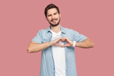 Photo for Portrait of bearded man in blue casual style shirt standing showing heart gesture over chest, being passionate, express love to close person. Indoor studio shot isolated on pink background. - Royalty Free Image