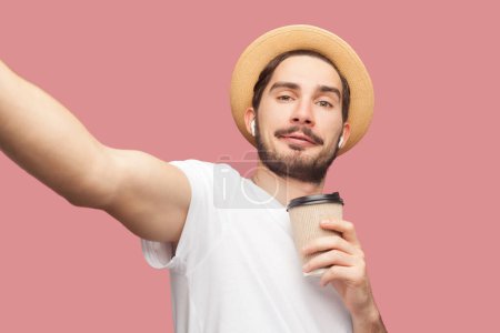 Photo for Portrait of smiling positive attractive bearded man blogger in white T-shirt and hat standing with coffee and making pointy of view photo. Indoor studio shot isolated on pink background. - Royalty Free Image