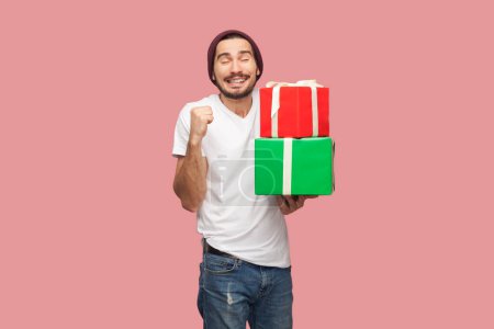 Photo for Portrait of satisfied extremely happy bearded man in white T-shirt and beany hat standing holding present box, clenched fist, celebrating. Indoor studio shot isolated on pink background. - Royalty Free Image