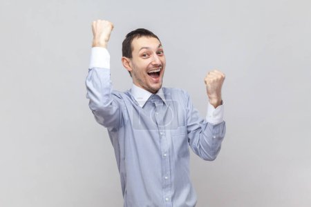 Photo for Portrait of overjoyed happy successful man standing with raised clenched fists, screaming with happiness, celebrating victory, wearing light blue shirt. Indoor studio shot isolated on gray background. - Royalty Free Image