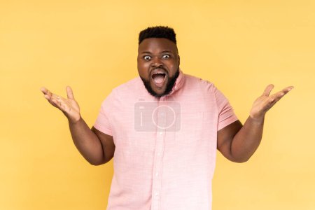 Photo for What a surprise. Portrait of man wearing pink shirt standing with wide open mouth and raising hands in amazement, shocked by unbelievable success. Indoor studio shot isolated on yellow background. - Royalty Free Image