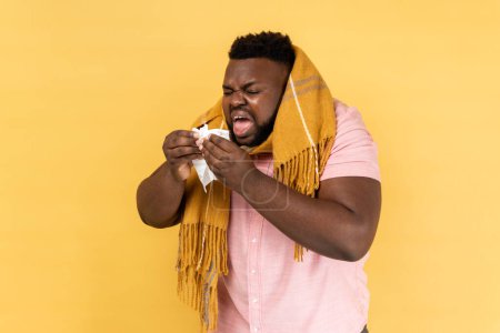 Photo for Portrait of unhealthy man in warm scarf coughing sneezing hard in napkin, feeling unwell with runny nose, severe seasonal allergy, influenza symptoms. Indoor studio shot isolated on yellow background. - Royalty Free Image