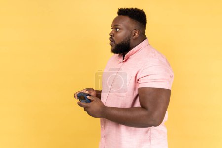 Photo for Side view of man wearing pink shirt holding in hands gamepad joystick, playing video games with big eyes and amazed facial expression. Indoor studio shot isolated on yellow background. - Royalty Free Image