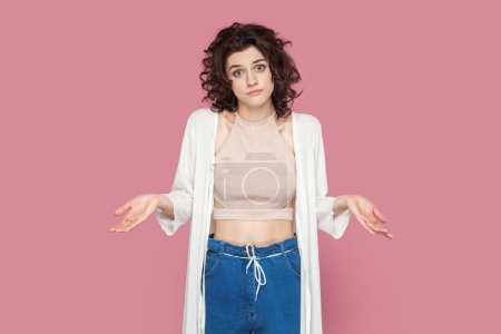 Photo for Portrait of puzzled uncertain woman with curly hairstyle wearing casual style outfit spreads hands aside, doesn't know answer. Indoor studio shot isolated on pink background. - Royalty Free Image