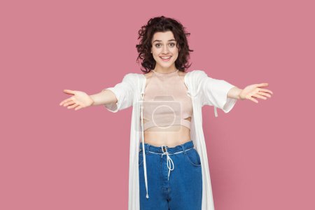 Photo for Welcome. Portrait of extremely happy pretty woman with curly hair wearing casual style outfit standing with outstretched hands, inviting. Indoor studio shot isolated on pink background. - Royalty Free Image