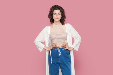 Photo for Portrait of self-confident egoistic woman with curly hair wearing casual style outfit standing with hands on hips, looking at camera, feels pride. Indoor studio shot isolated on pink background. - Royalty Free Image