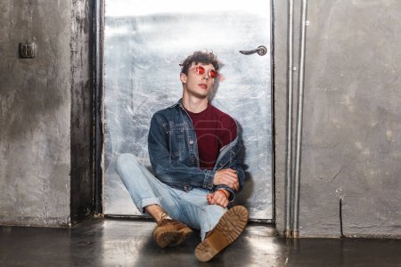 Photo for Full length portrait of calm attractive relaxed fashionable model man in bright red sunglasses and denim casual style, sitting near metallic door, looking at camera. Indoor studio shot. - Royalty Free Image