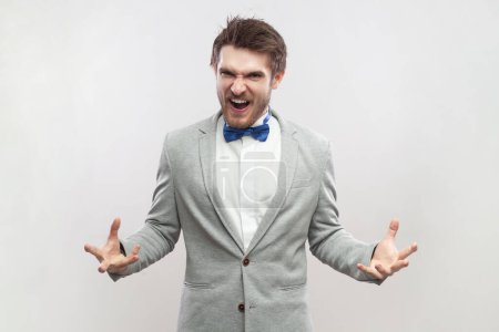 Photo for Portrait of angry aggressive handsome bearded man standing with raised arms, screaming, expressing hate and anger, wearing grey suit and blue bow tie. Indoor studio shot isolated on gray background. - Royalty Free Image